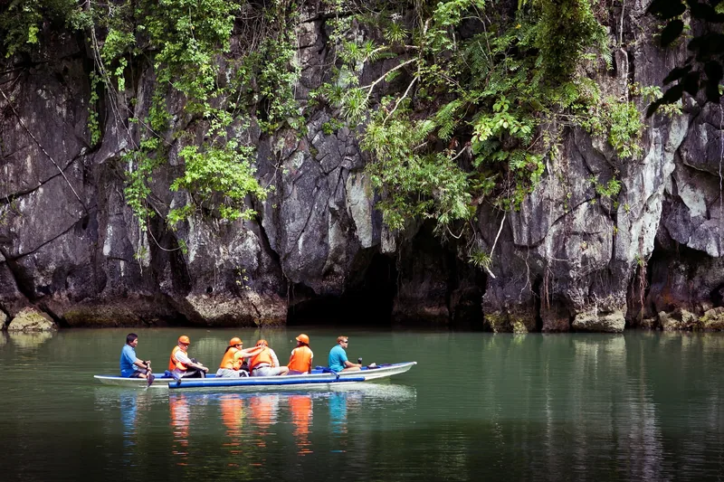 A small outrigger boat carries tourists in life vests toward a cave mouth beneath limestone cliffs in Palawan.