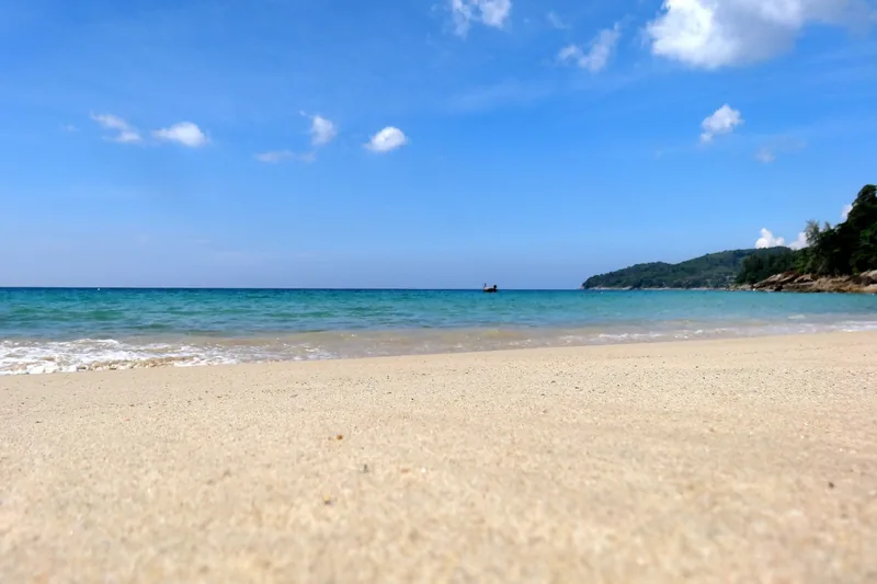 A quiet sandy beach in Phuket with turquoise water and a distant boat under a bright blue sky.