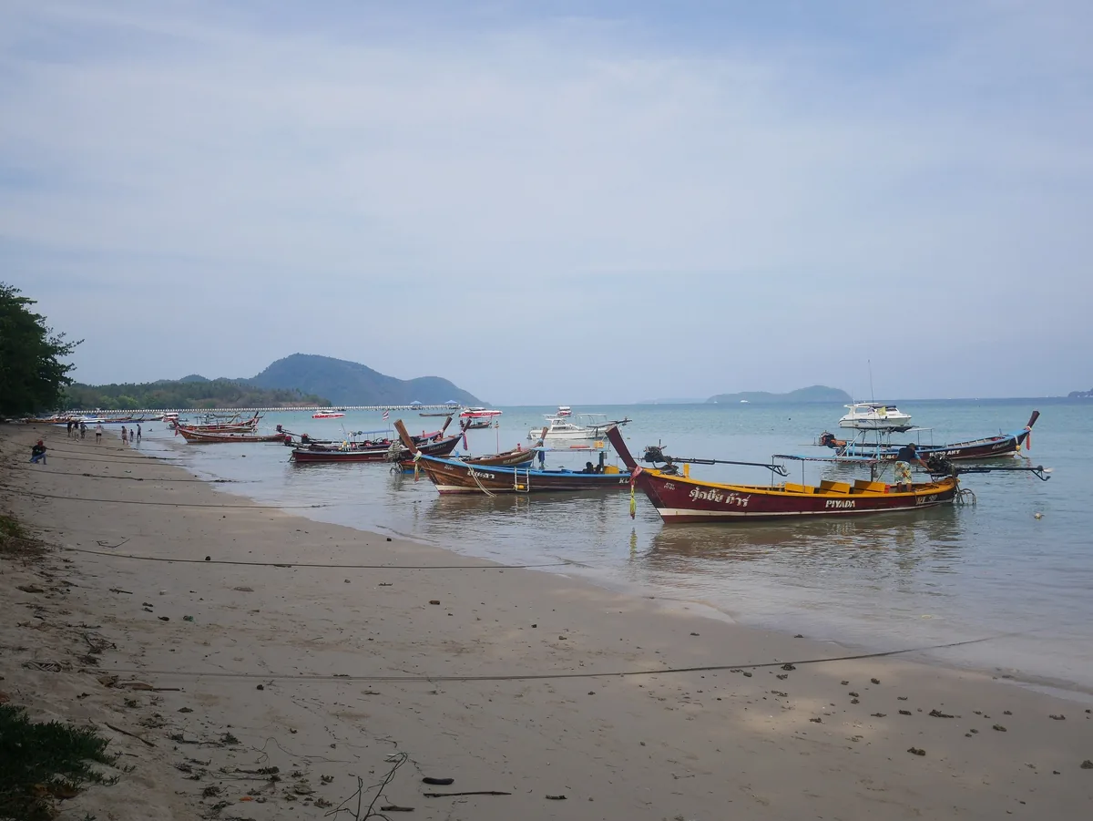 Longtail boats moored along the sandy shore of Rawai Beach in Phuket with hills in the distance.