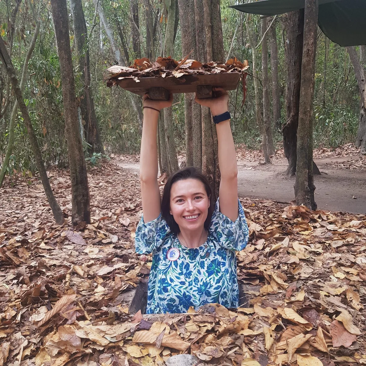 A smiling tourist emerges from a hidden trapdoor entrance at the Cu Chi Tunnels near Saigon, lifting the leaf-covered cover.