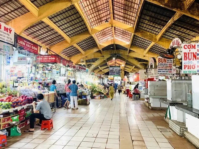 Shoppers stroll the tiled interior aisle of a covered market in Saigon with food stalls and signboards.