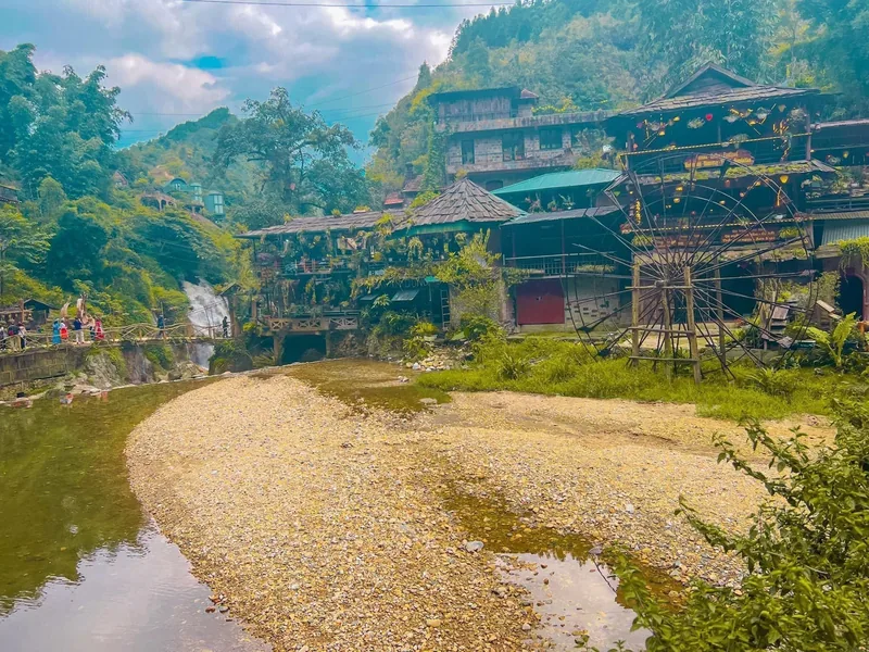Wooden stilt houses and a waterwheel line a river with a small waterfall at Cat Cat Village in Lao Cai.