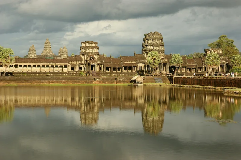 Angkor Wat's stone towers and galleries reflect in the surrounding moat under a cloudy sky in Siem Reap.