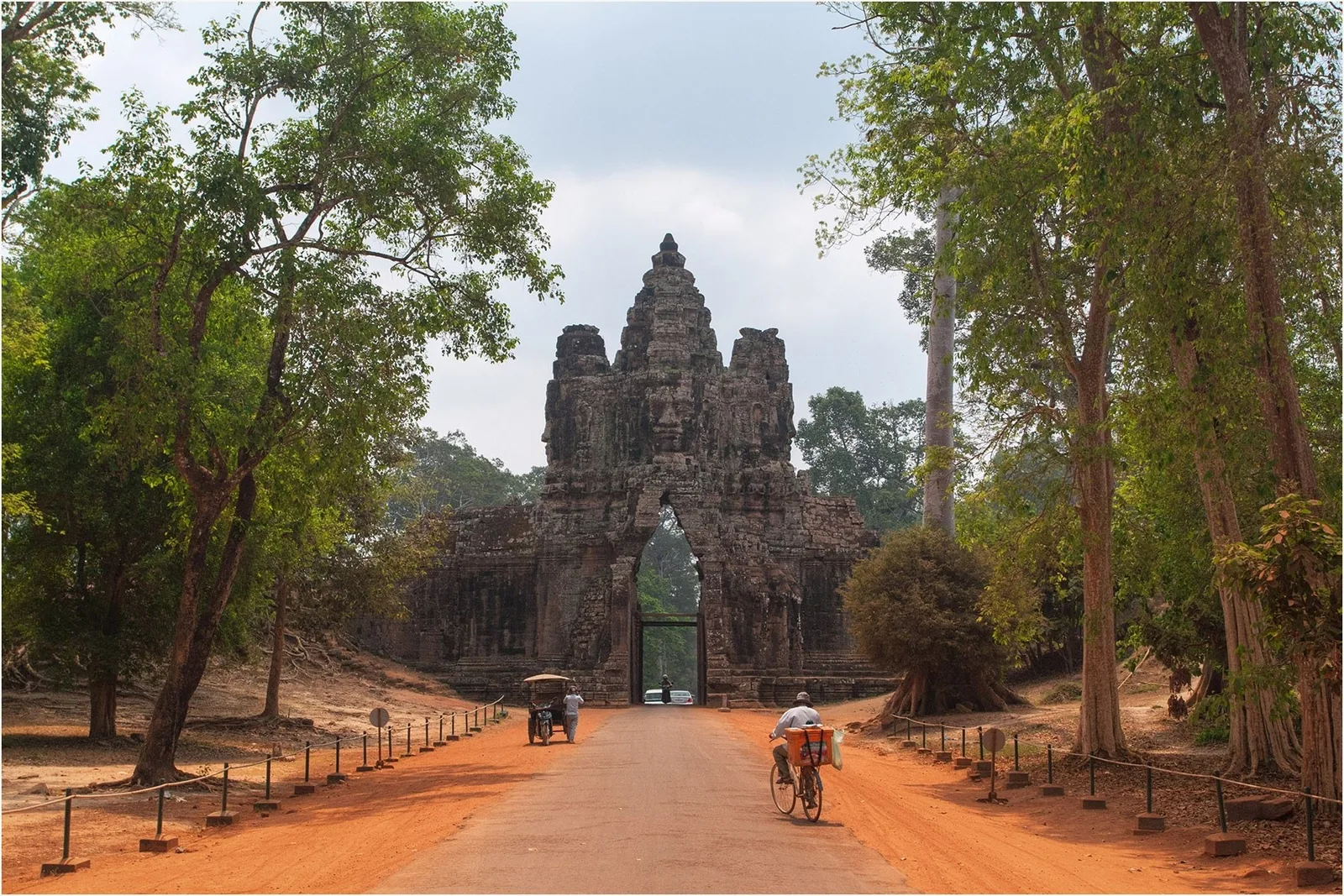 A stone gate tower of Angkor Thom rises at the end of a red dirt road lined with tall trees in Siem Reap.