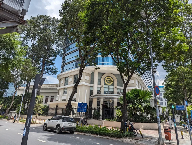 A tree-lined street in District 1 leads to a modern glass tower rising above a classical white building in Ho Chi Minh City.