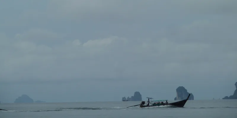 A long-tail boat carries passengers across calm sea past limestone karst islands near Krabi, Thailand.