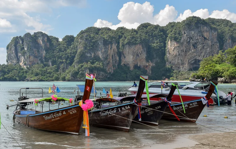 Traditional longtail boats decorated with ribbons rest on the beach beneath limestone cliffs at Railay, Krabi.