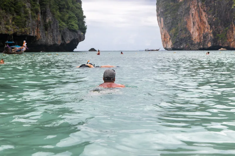 Swimmers glide through clear turquoise water between limestone cliffs near Krabi, Thailand.