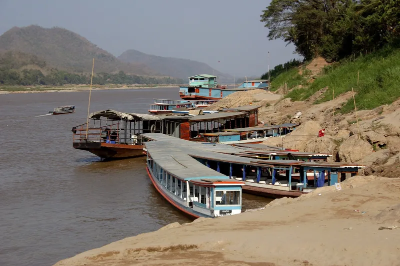 Wooden riverboats moored along the muddy bank of the Mekong River near Luang Prabang, with hills in the distance.