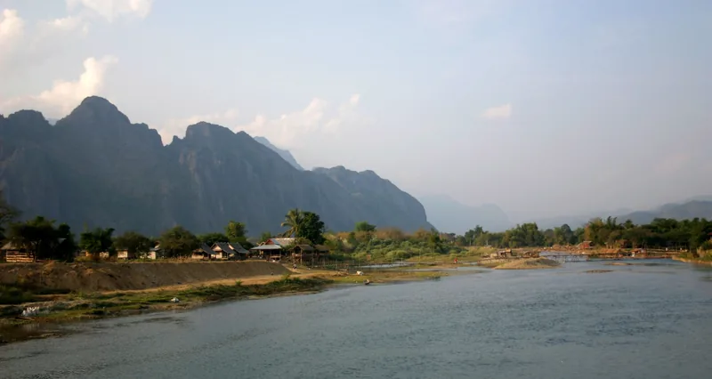 Karst limestone mountains rise above the Nam Song River and riverside huts in Vang Vieng, Laos.
