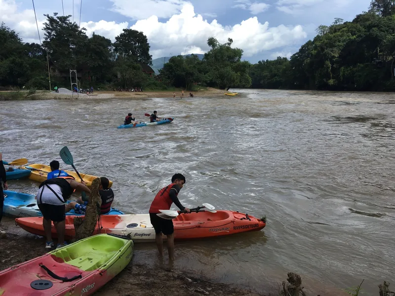 Kayakers prepare colorful kayaks on the muddy Nam Song River in Vang Vieng, surrounded by karst hills.