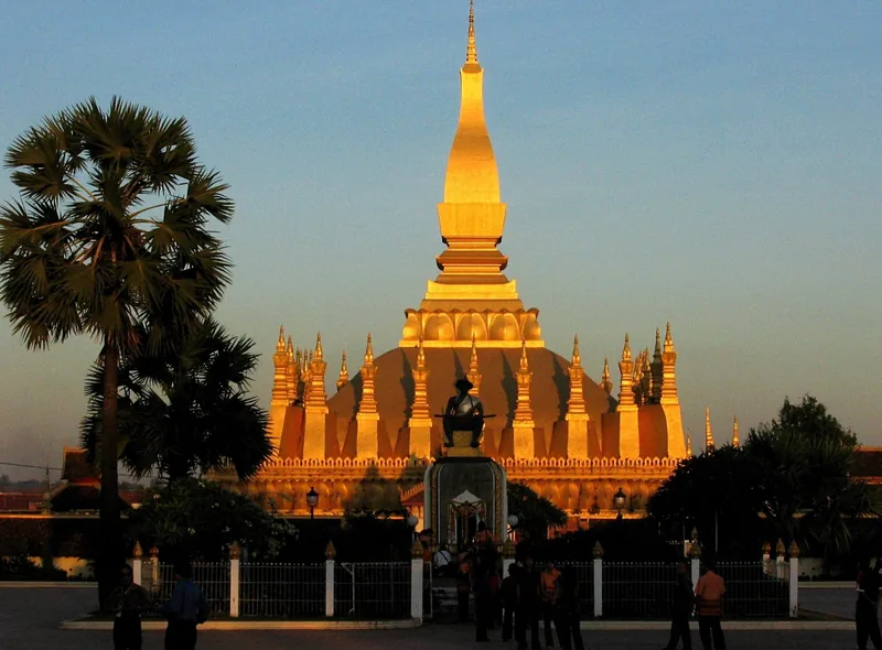 Golden spires of Pha That Luang stupa glow at sunset in Vientiane, framed by a palm tree.