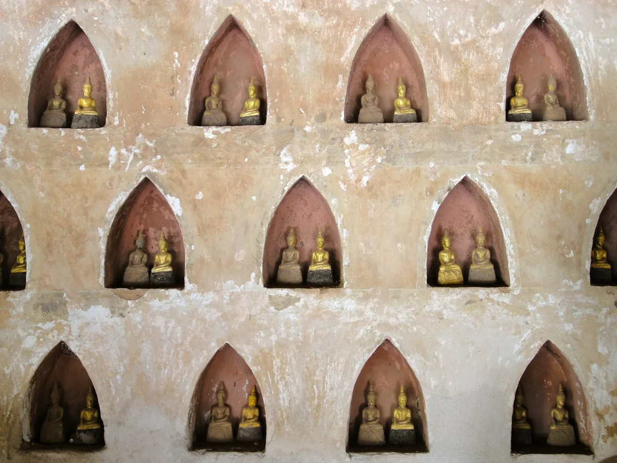 Rows of small gilded Buddha statues rest in arched niches on a weathered temple wall in Vientiane, Laos.
