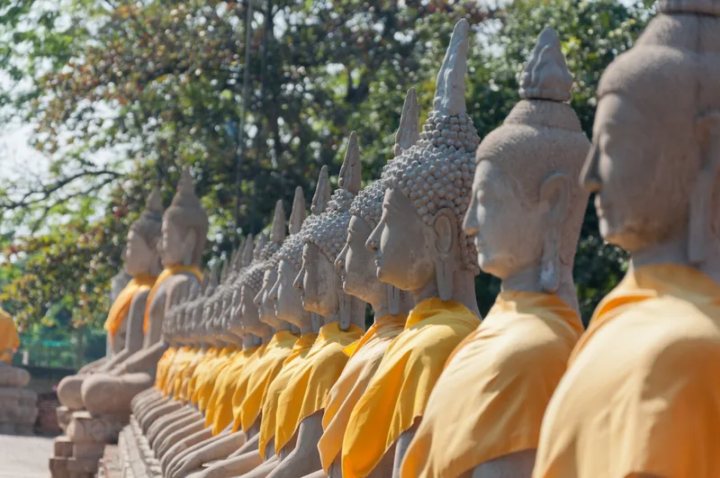 A long row of stone Buddha statues draped in orange sashes at a temple in Ayutthaya, Thailand.