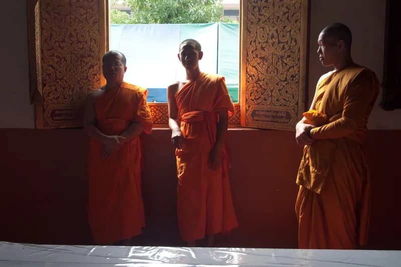 Three Buddhist monks in orange robes stand by carved gilded windows inside a temple in Chiang Mai.