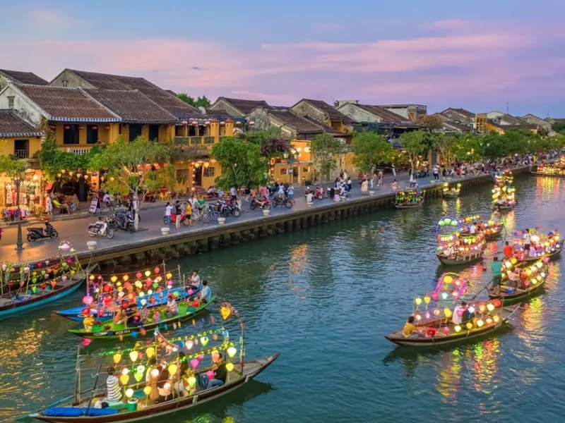 Lantern-lit boats drift along Hoi An's river at dusk beside the town's glowing old quarter buildings.