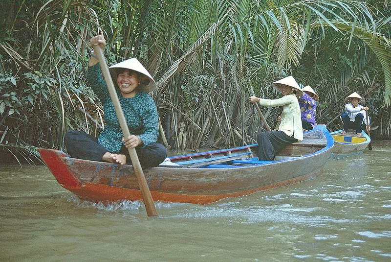 Smiling women in conical hats row wooden sampans through a narrow palm-lined canal in the Mekong Delta.