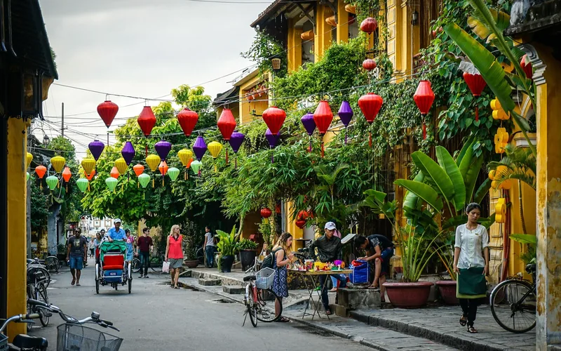Colorful silk lanterns hang above a lively street lined with yellow buildings in Hoi An's old town.