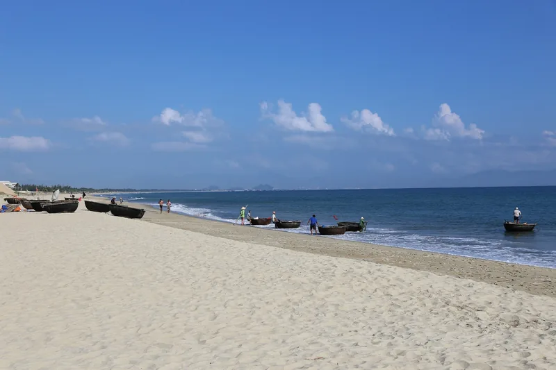 Round basket boats rest on a sandy beach near Hoi An under a blue sky with scattered clouds.