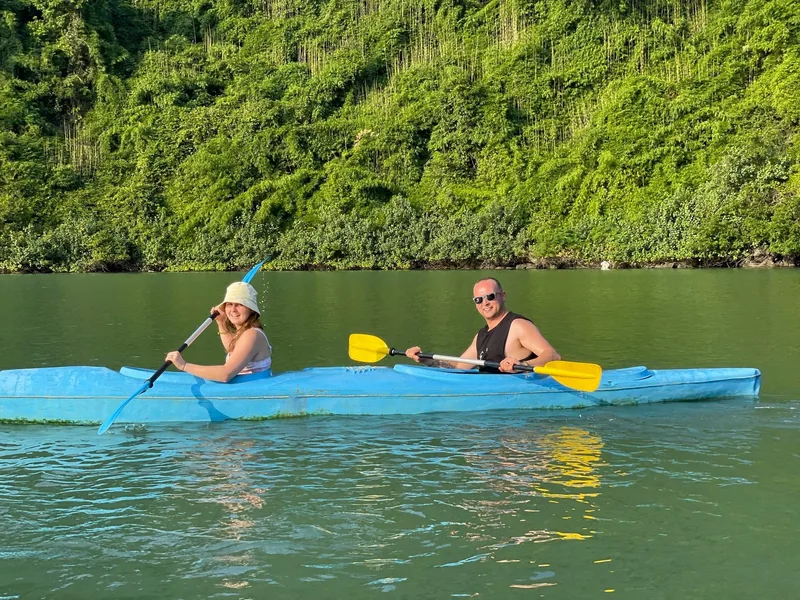 A couple paddles a blue kayak along calm green water beside forested cliffs in Lan Ha Bay.