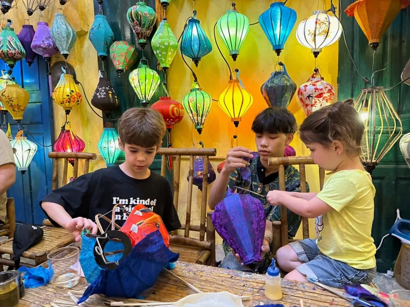 Children learn to make colorful silk lanterns at a workshop surrounded by hanging lanterns in Hoi An.