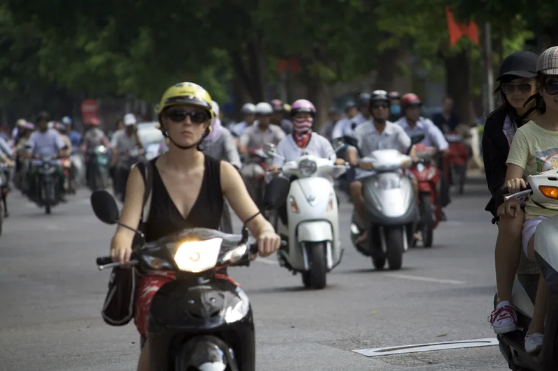 Riders in helmets navigate a busy motorbike traffic stream on a tree-lined street in Hanoi.