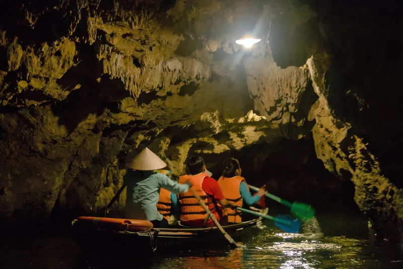A boatwoman rows tourists in life jackets through a dimly lit limestone cave at Trang An, Ninh Binh.
