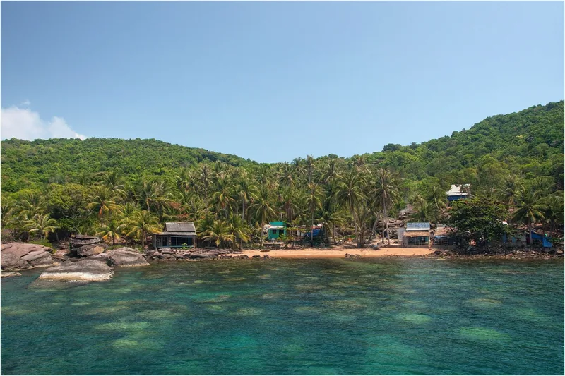 Small huts sit among palm trees along a rocky shore with clear turquoise water near Phu Quoc's An Thoi archipelago.