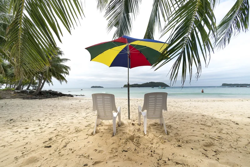 Two beach chairs sit under a colorful umbrella framed by palm leaves on a sandy Phu Quoc beach.